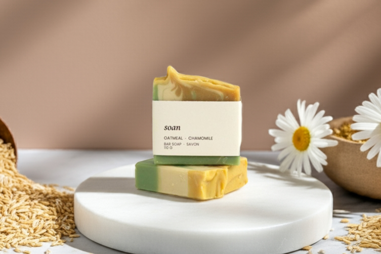 Two oatmeal chamomile bars of soap on a marble surface with seeds and flowers, against a neutral background.