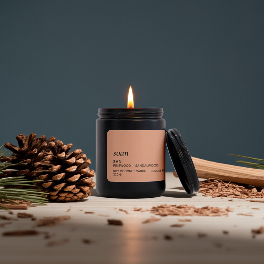Lit San Pinewood Sandalwood Soy candle in a black container with a label, surrounded by pine cones and wood on a dark background