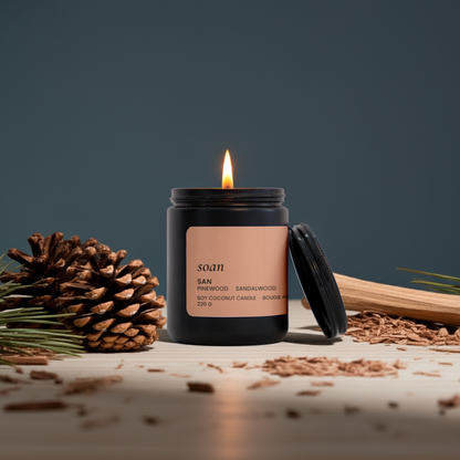 Lit San Pinewood Sandalwood Soy candle in a black container with a label, surrounded by pine cones and wood on a dark background