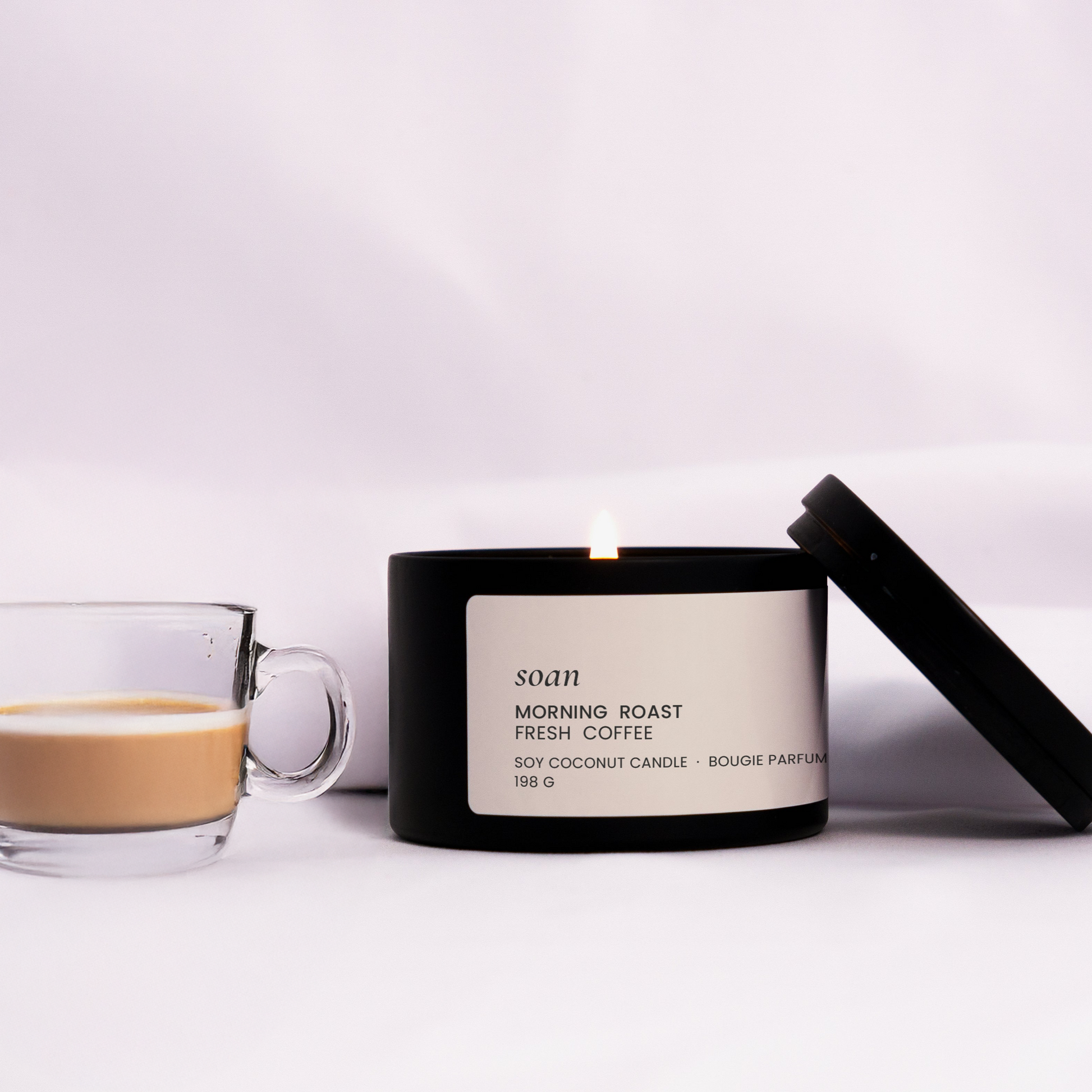 Candle labeled 'Morning Roast Frensh Coffee' next to a glass of coffee on a white surface.