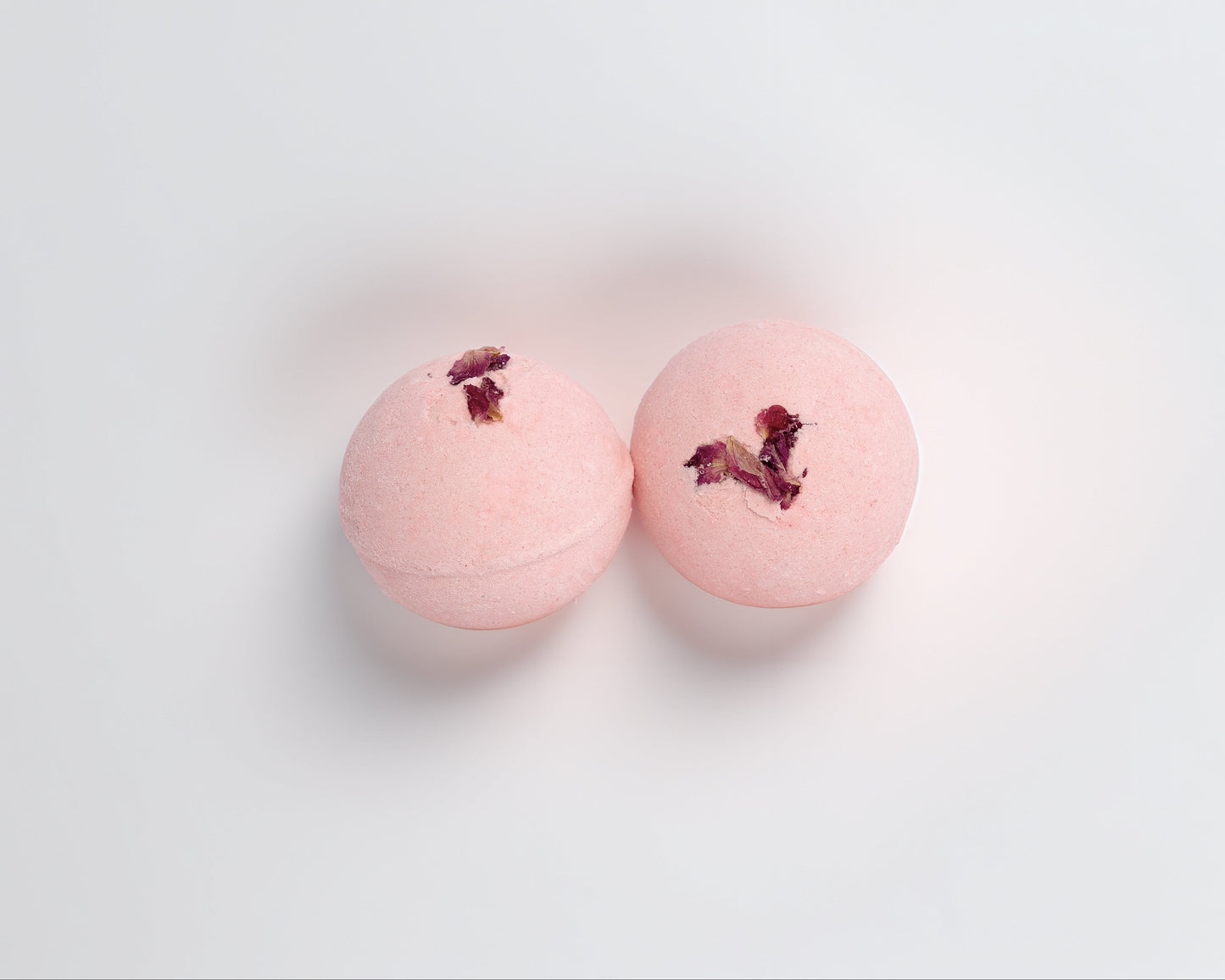 Two pink bath bombs with rose scent with rose petals on a light gray background