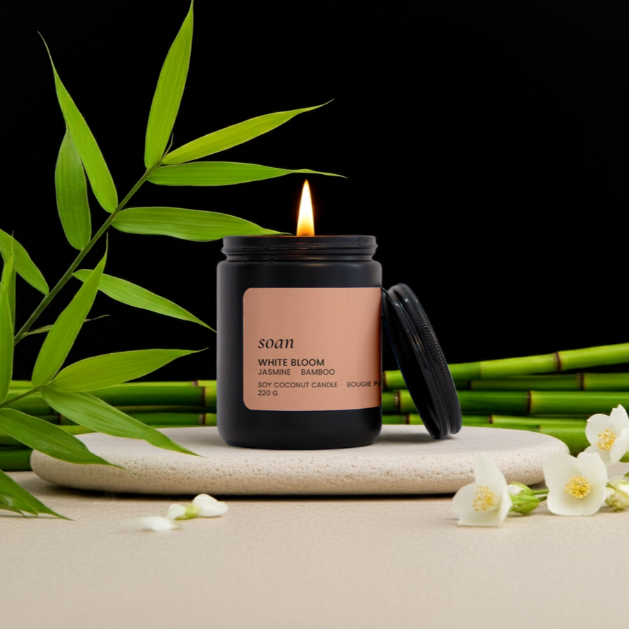 White Bloom Black candle with a beige label on a stone surface with bamboo and flowers, against a black background.