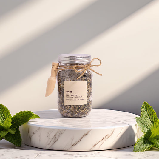 Jar of bath salts with a label on a marble surface, surrounded by mint leaves.