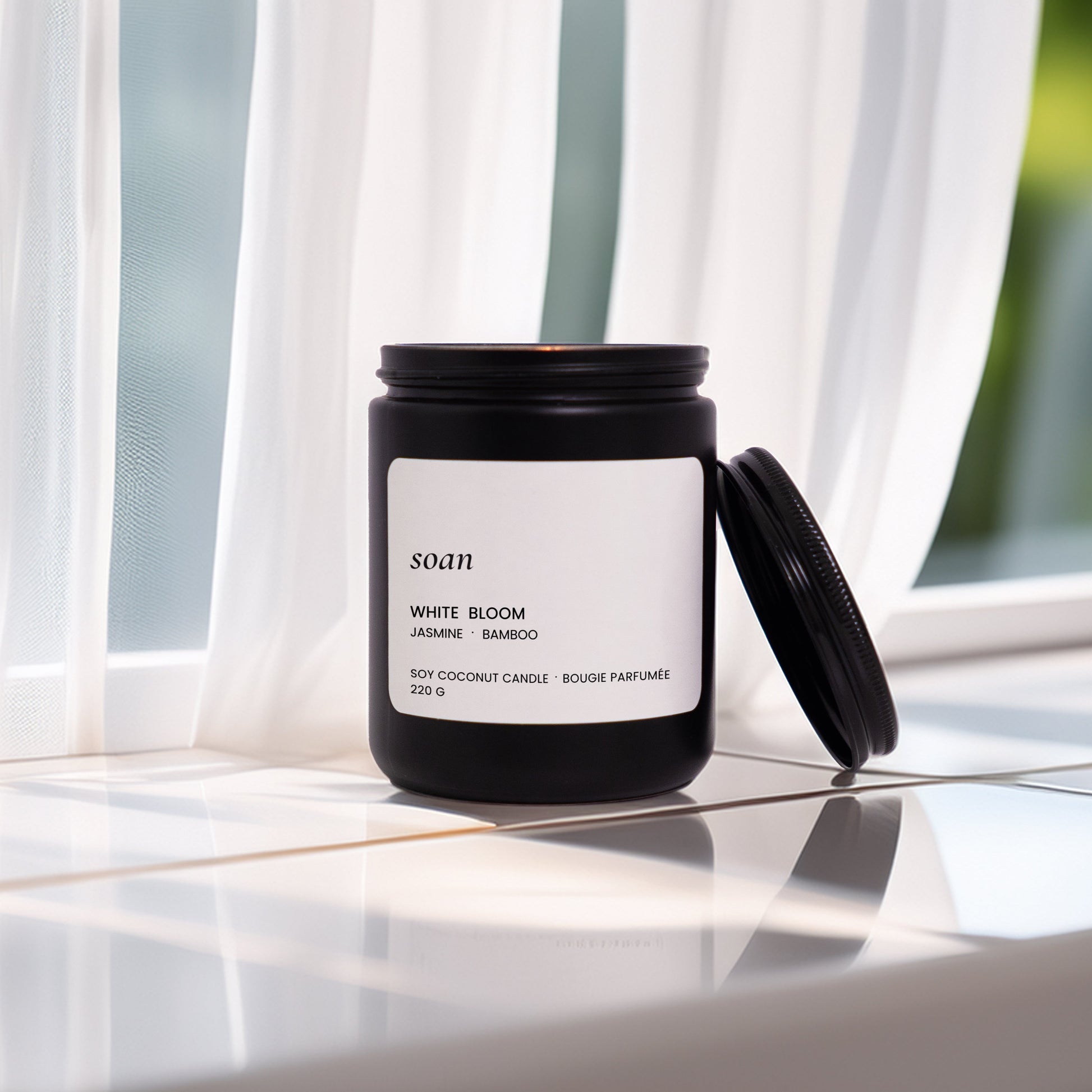 Black candle with a white label on a reflective surface with curtains in the background