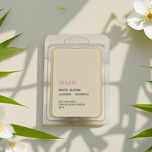 Packaged wax meltwith 'White Bloom' fragrance surrounded by white flowers and green leaves on a light background